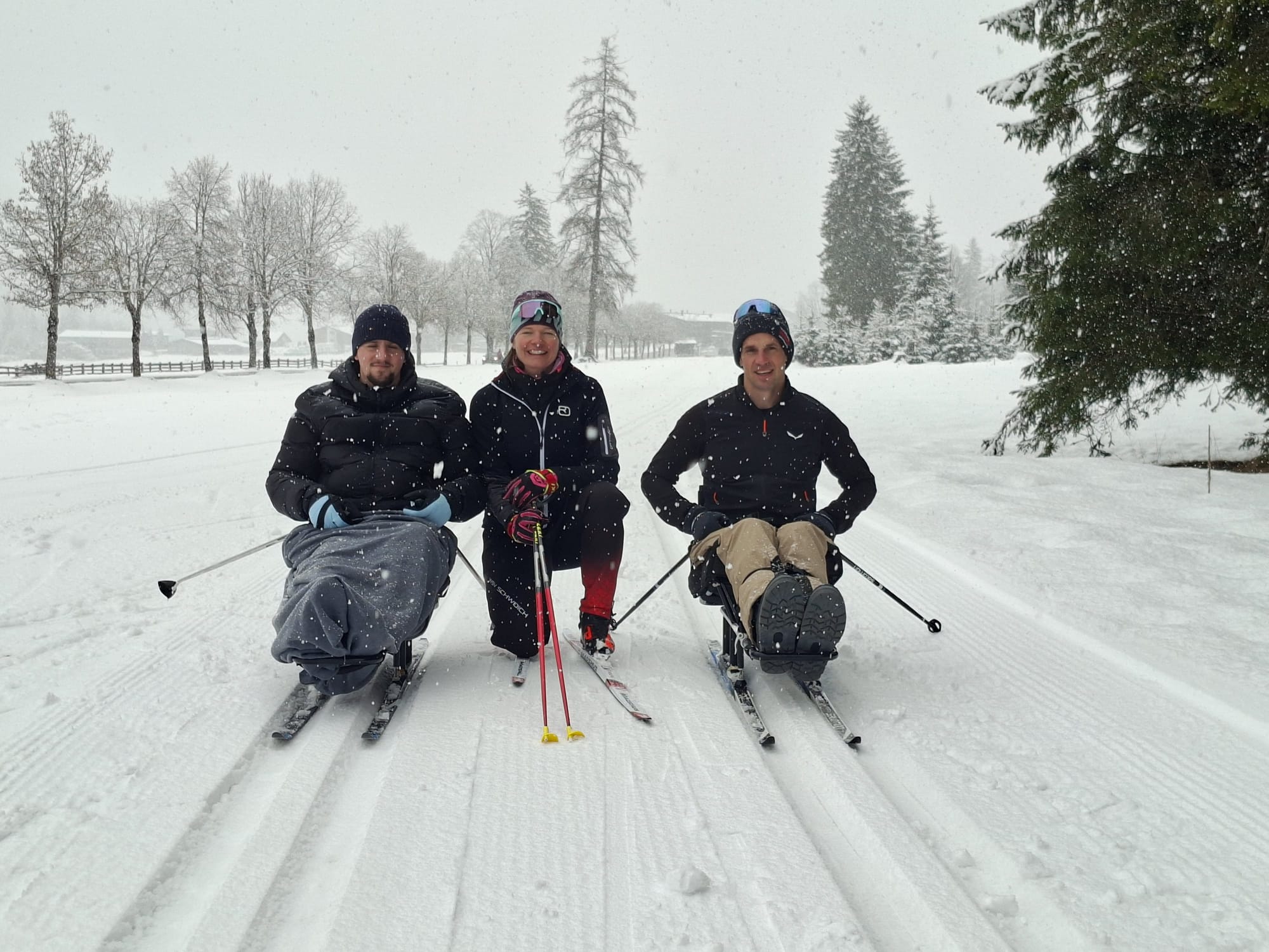 Zwei Teilnehmende auf Langlaufschlitten im Schnee, in der Mitte zwischen ihnen ist die Trainerin zu sehen