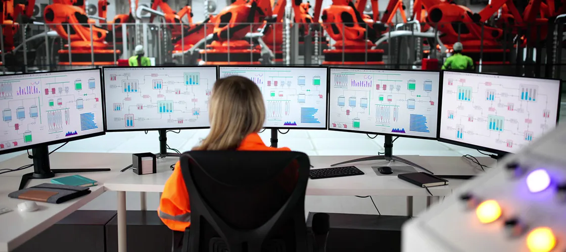 Adobestock 1451505302 ©Andrey Popov Eine Frau sitzt vor fünf Monitoren, die im Halbkreis vor ihr angeordnet sind. Im Hintergrund sieht man Industrieroboter