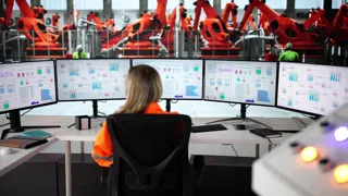 Adobestock 1451505302 ©Andrey Popov Eine Frau sitzt vor fünf Monitoren, die im Halbkreis vor ihr angeordnet sind. Im Hintergrund sieht man Industrieroboter