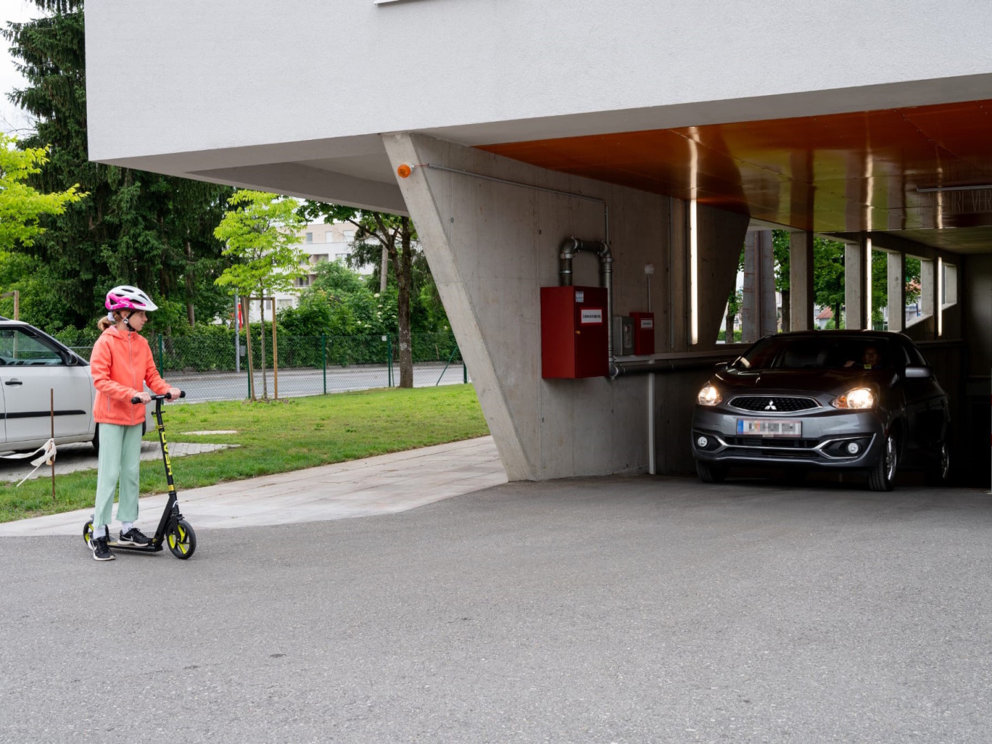 Ein Mädchen steht auf einem Tretroller vor einer Garagenausfahrt, aus der ein Pkw kommt.