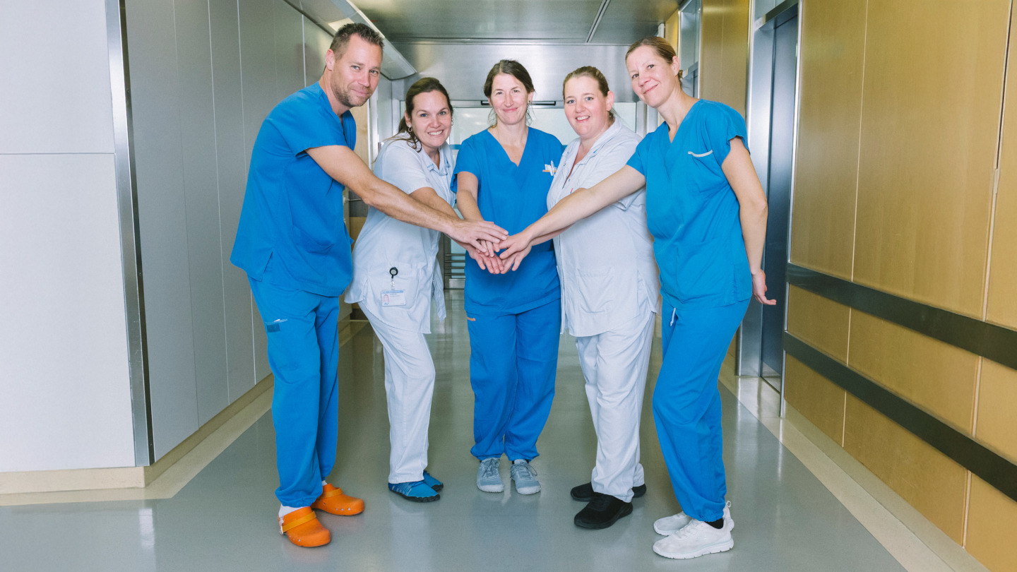 Gruppenfoto, auf dem fünf Mitglieder des Flex Teams Pflege Ost in einem Krankenhausgang stehen und die ihre Hände als Zeichen des Teamzusammenhalts aufeinander legen. 