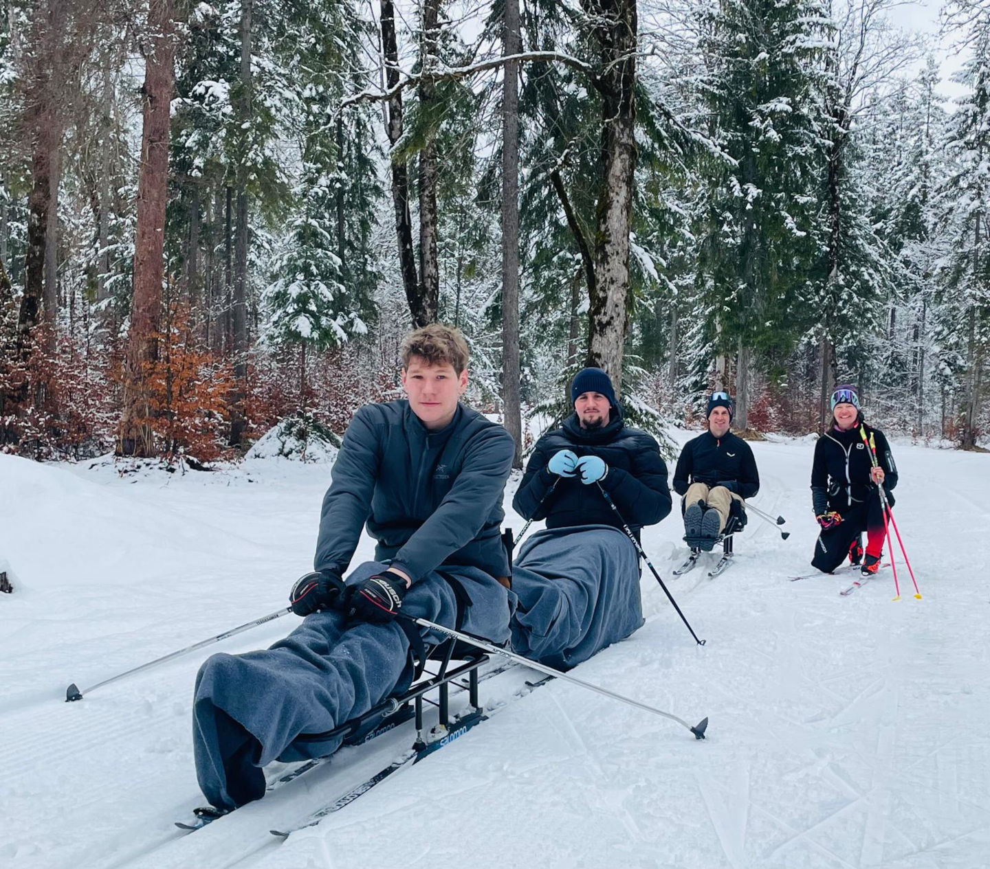Drei Teilnehmende auf Langlaufschlitten im Schnee, rechts im Bild ist die Trainerin zu sehen