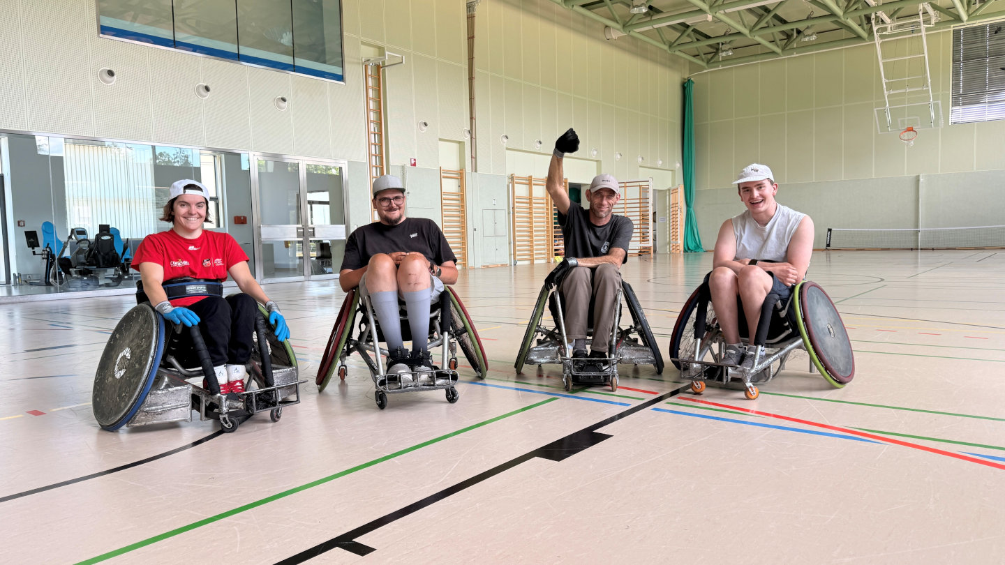 Vier Rollstuhl-Rugby-Spieler:innen befinden sich in einem Turnsaal und blicken zum Fotografen